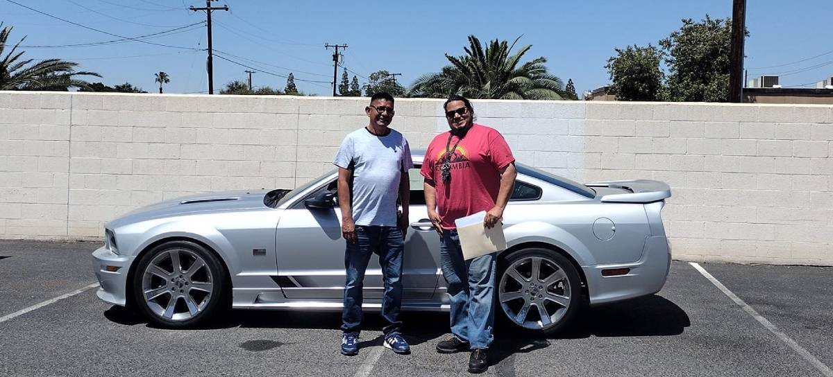 Side view of silver muscle car with 2 people standing in front of it